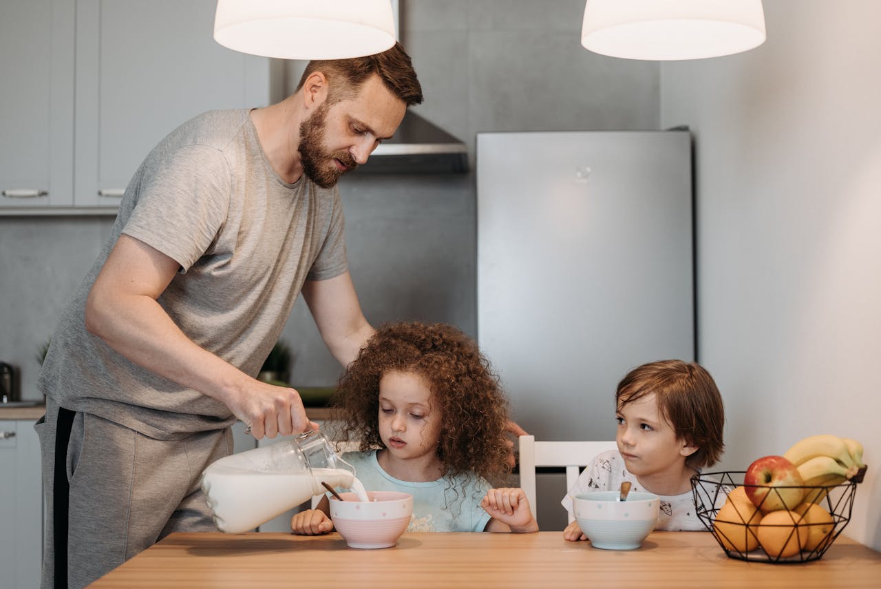 A father pours milk into cereal bowls for his children at the breakfast table in a modern kitchen.